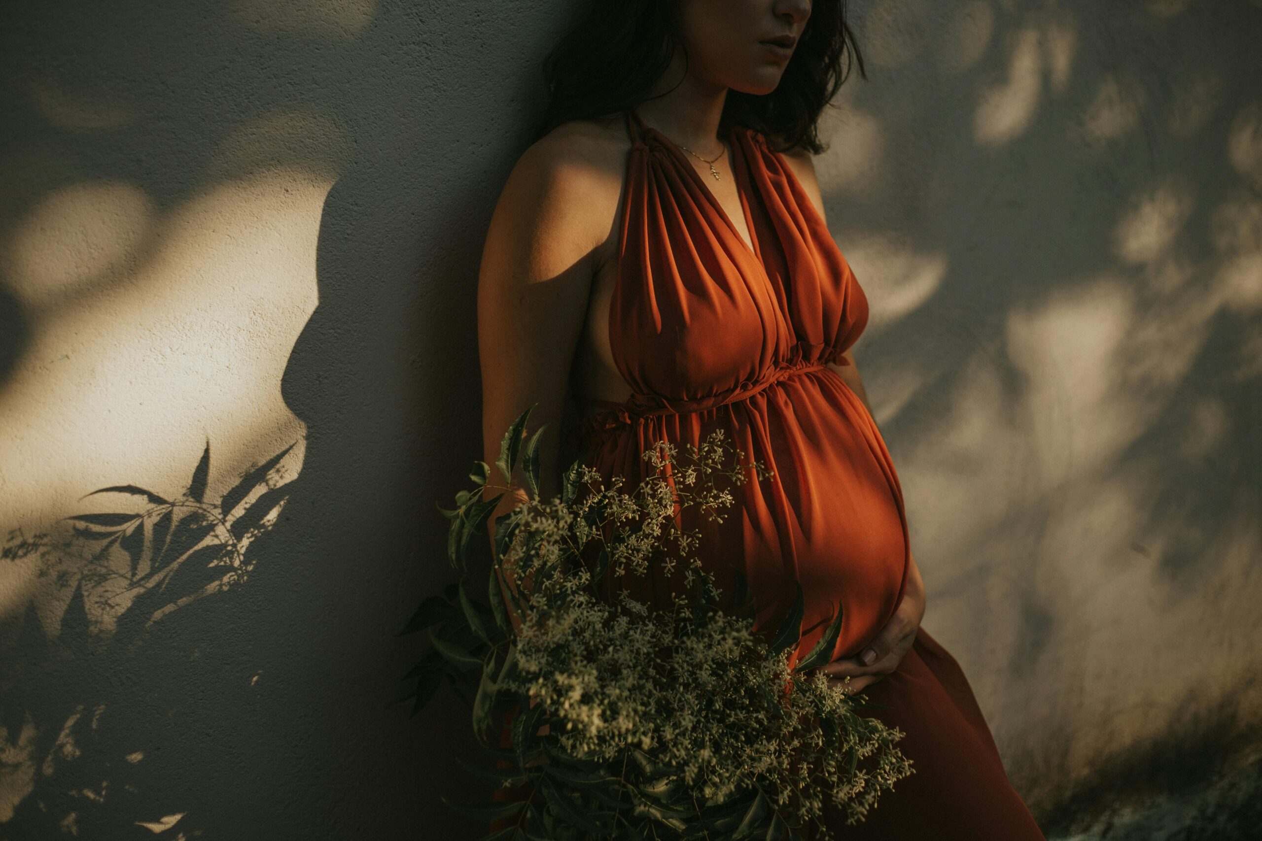 A serene pregnant woman in a vibrant dress holds flowers against a textured wall.